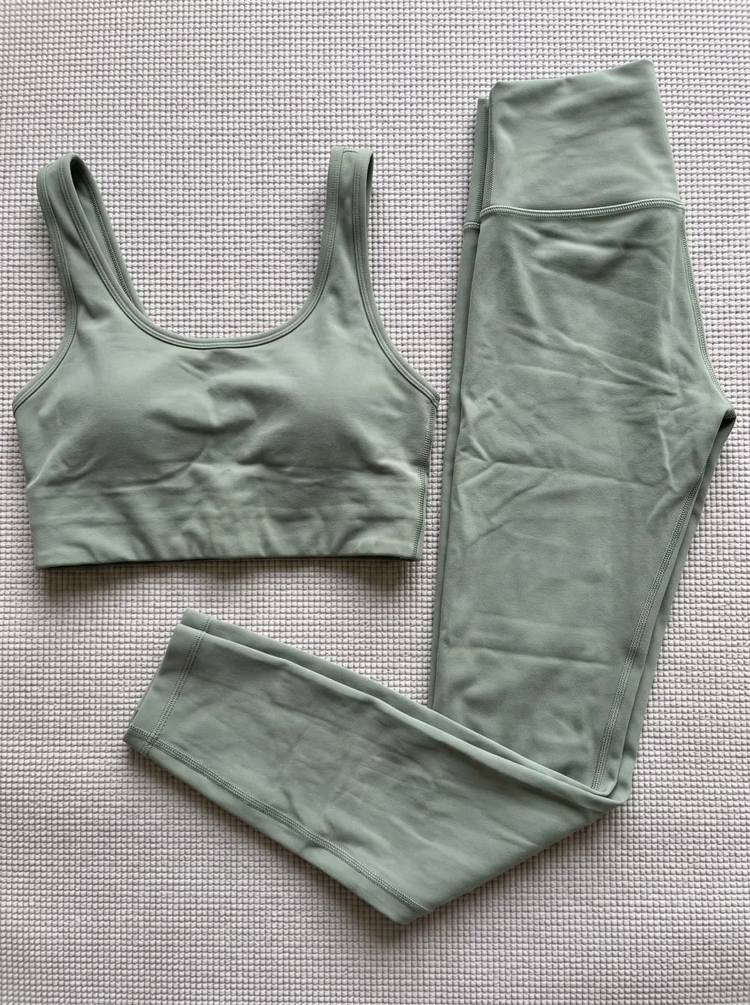 A sage sports bra and matching leggings laid flat side by side on a woven mat