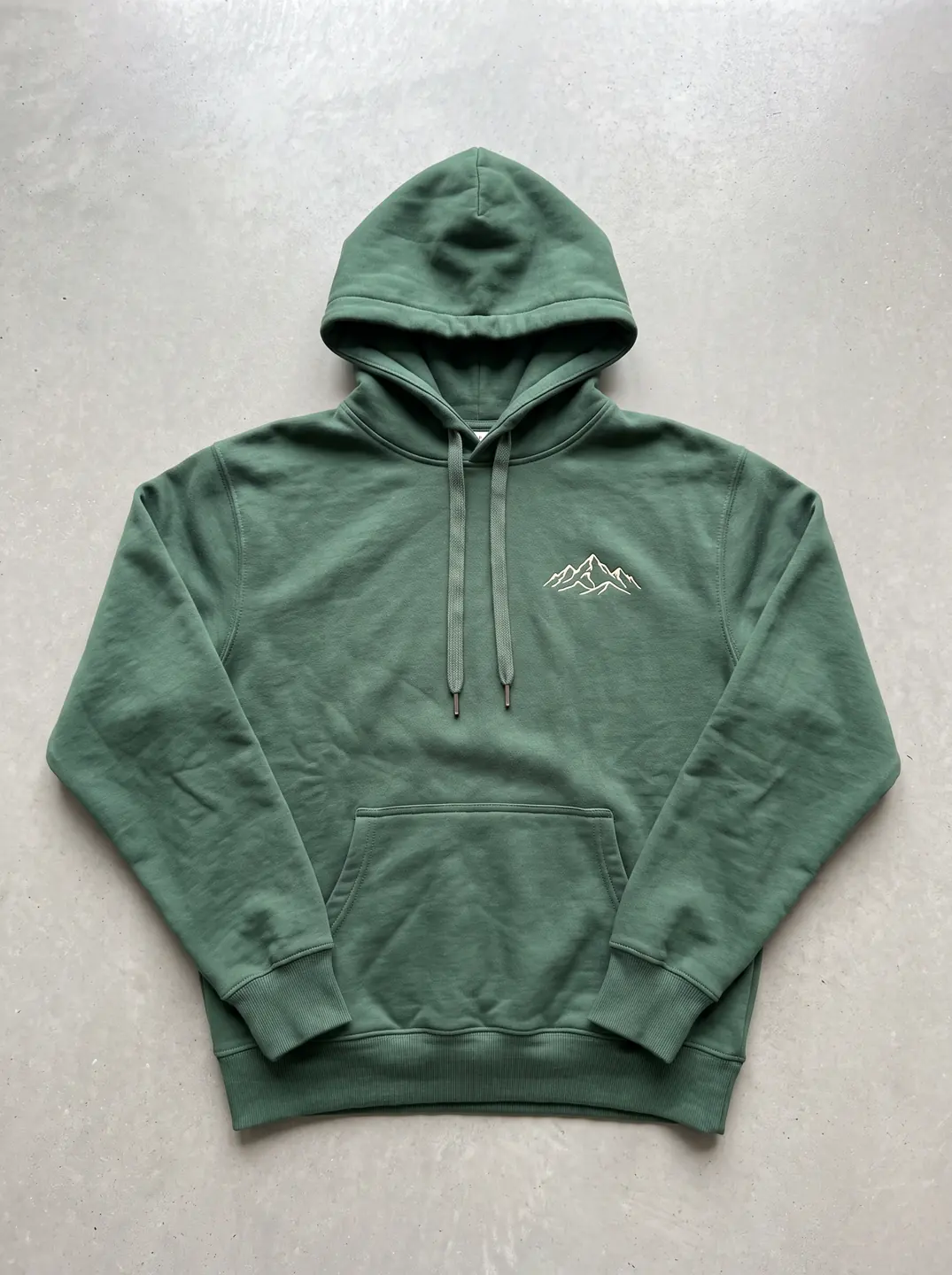 A green hoodie with a small white mountain emblem laid flat on pale concrete