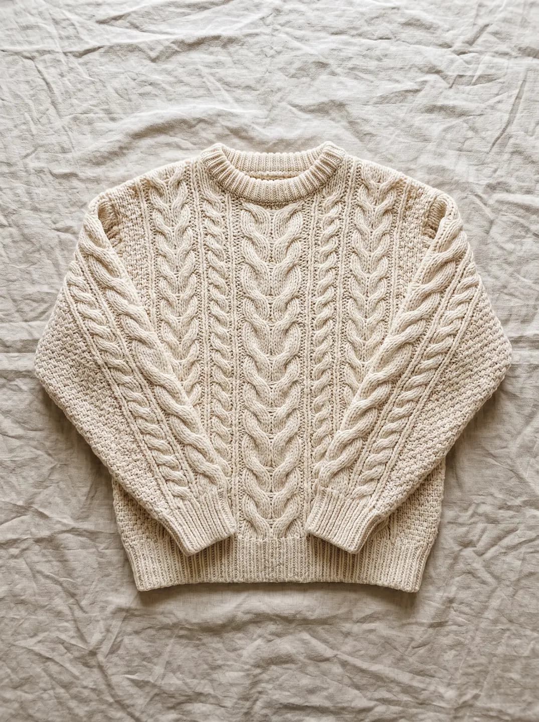 A cream cable-knit sweater laid flat on crumpled natural linen