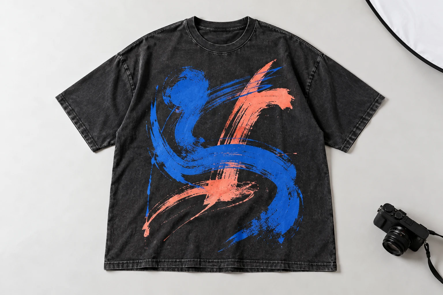 A complete washed-black graphic T-shirt laid flat beside a camera on a clear studio surface.