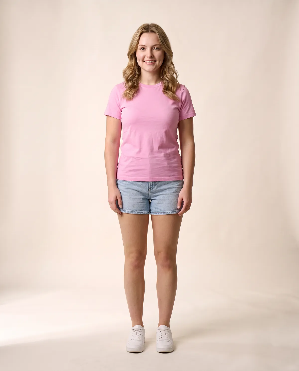 A model wearing the pink T-shirt with light denim shorts and white trainers, full length in a warm studio
