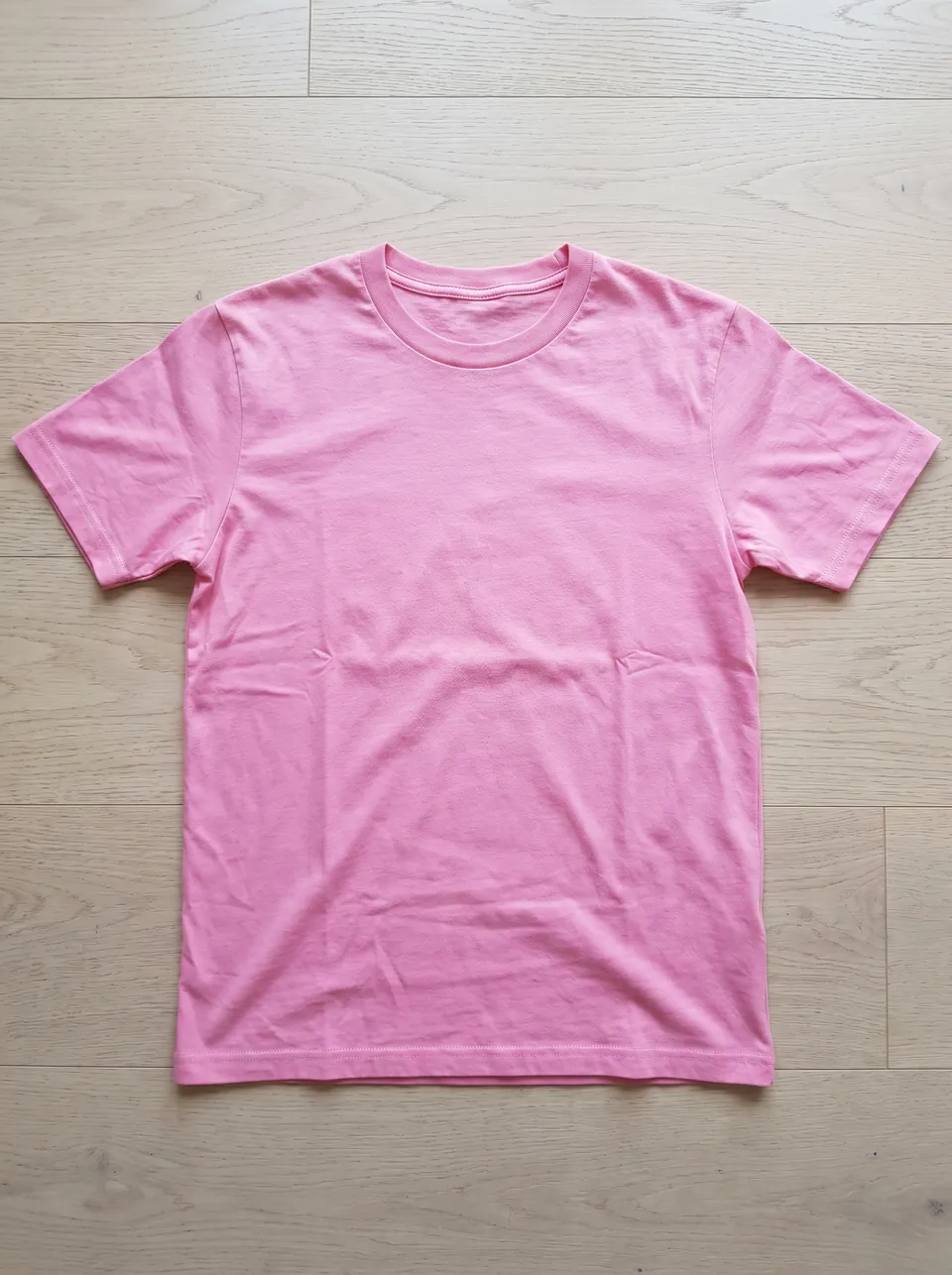Phone photo of a plain pink crew-neck T-shirt laid flat on a pale wooden floor